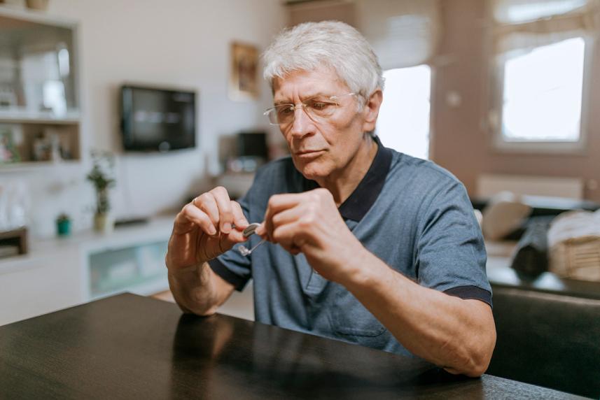 Man inspecting a hearing aid, sitting at a table in a home