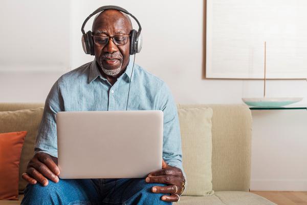 A man sitting on a couch, holding and looking at a laptop, with headphones on